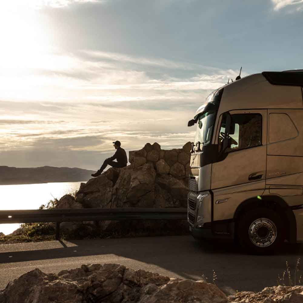 A driver sits on a rock looking out to sea at sunset, with his Volvo FH truck parked beside him